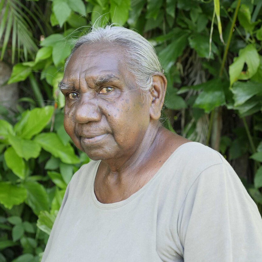 Older indigenous woman smiling and looking forwards with body side on, wearing grey shirt in front of leafy background