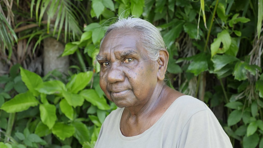 Older indigenous woman smiling and looking forwards with body side on, wearing grey shirt in front of leafy background