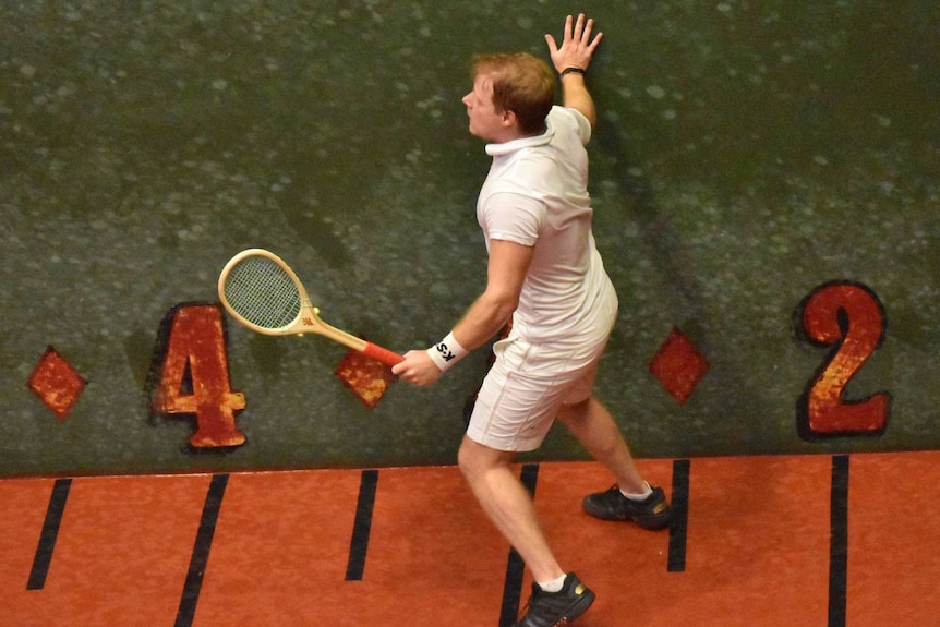 A man with a wooden racquet leans against a real tennis court wall