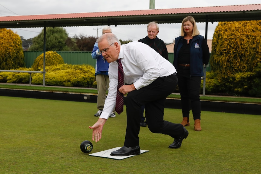 Scott Morrison bowls ball along the ground as people watch on.