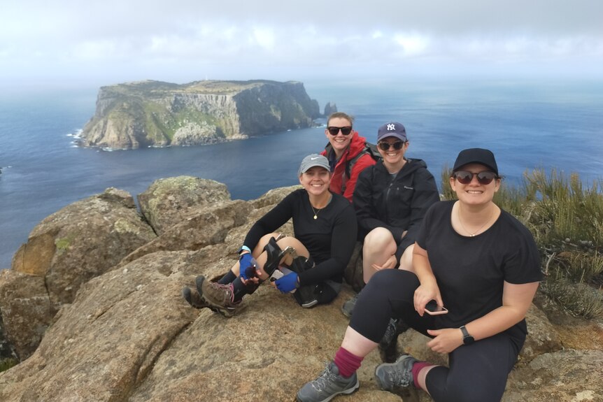 Four bushwalkers lounge on a high rock platform looking over a spectacular coastal island