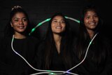 Three smiling girls sitting against a black wall, with a glowing skipping rope draped around them.