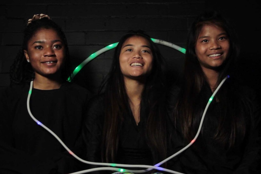 Three smiling girls sitting against a black wall, with a glowing skipping rope draped around them.