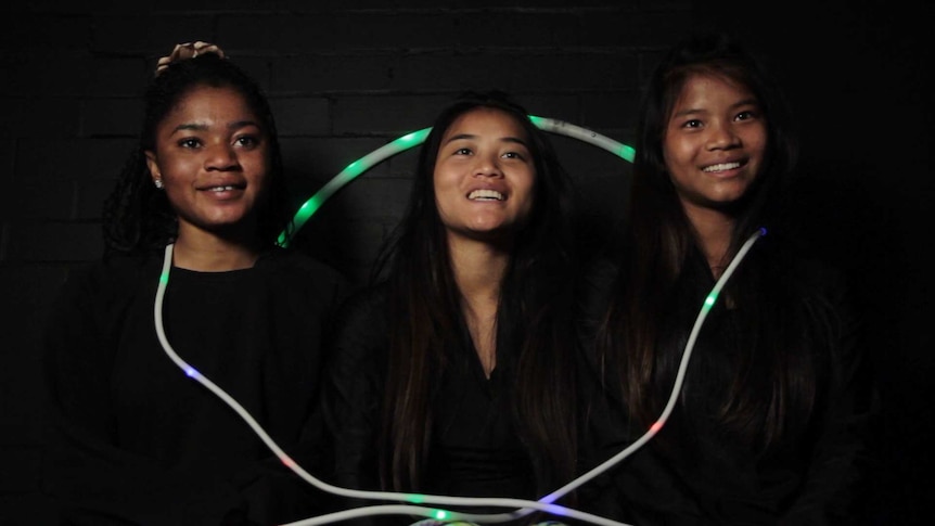 Three smiling girls sitting against a black wall, with a glowing skipping rope draped around them.