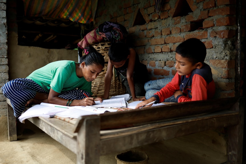 A woman and two children on a wooden bed in a brick room, working on school work 