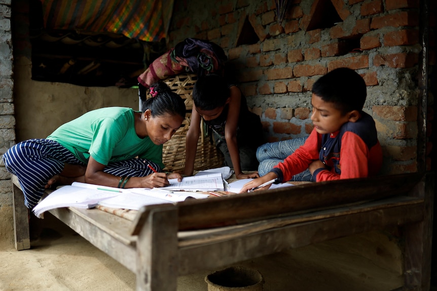 A woman and two boys on a wood bed in a brick room, working on school work 