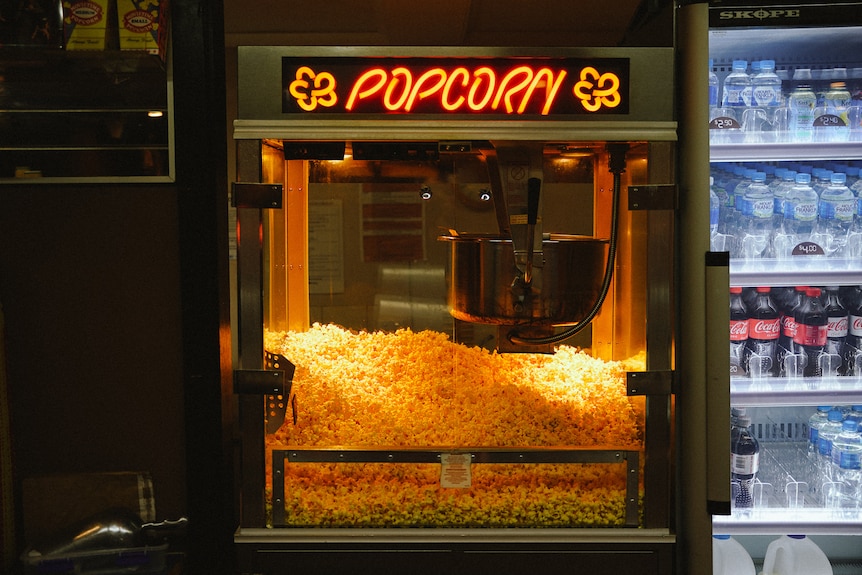 A large popcorn warmer with popcorn inside and the word 'popcorn' lit up in yellow neon signage.