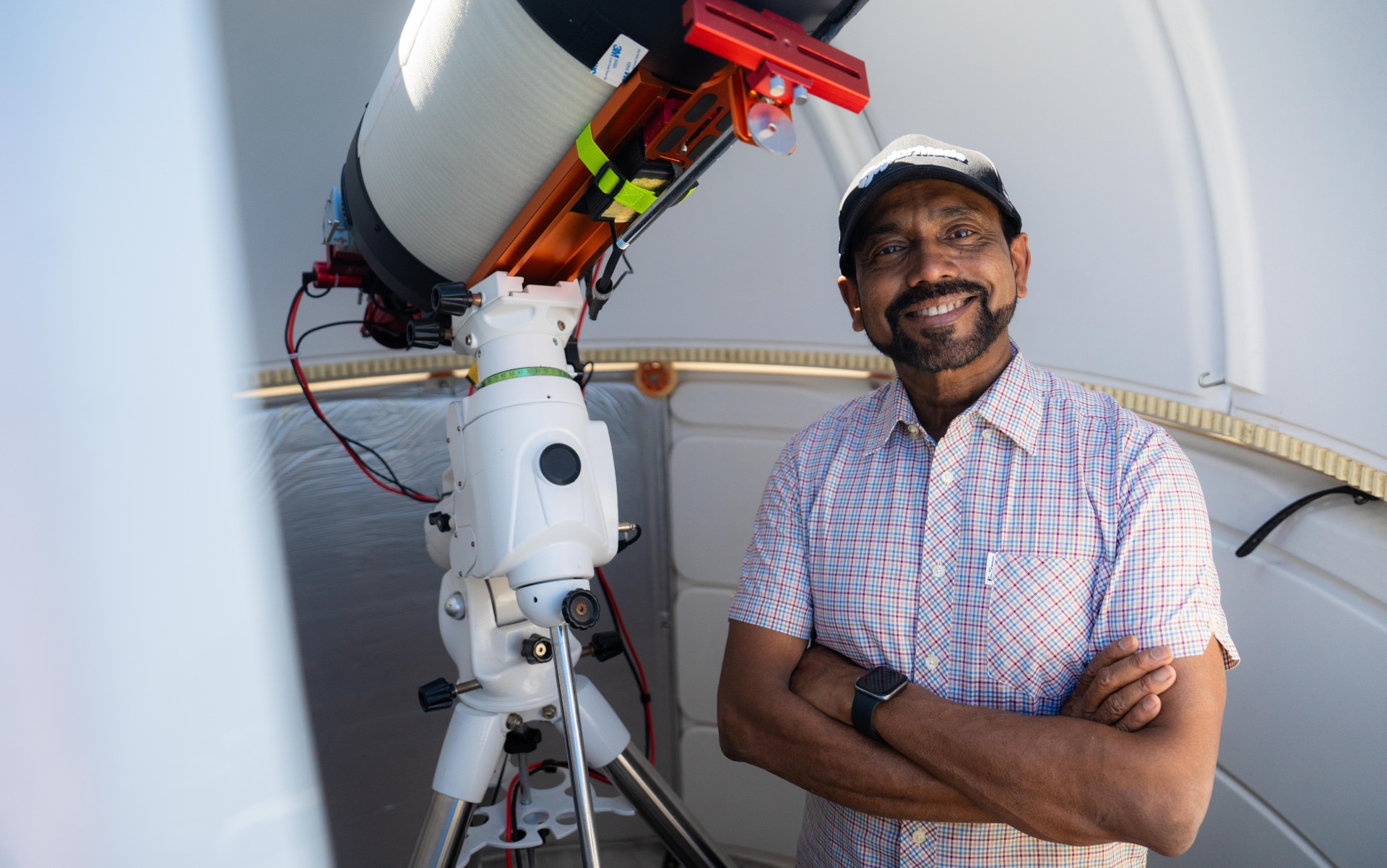 A man wearing a cap smiling and standing next to a telescope.