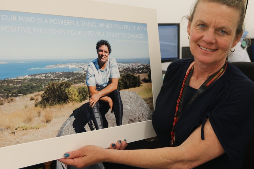 Woman on right holding up a large photograph on the right of a man perched on a rock on a hill.