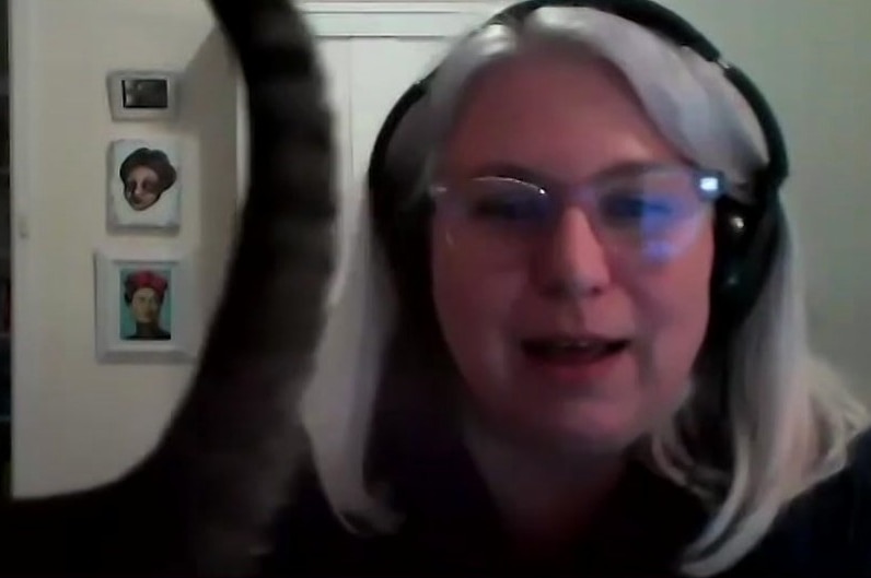 A woman wearing glasses and headphones on a video chat as her cat walks past.