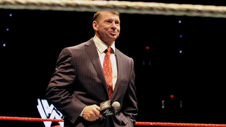 Vince McMahon in a suit and tie holding a microphone inside a wrestling ring with the WWE logo behind him