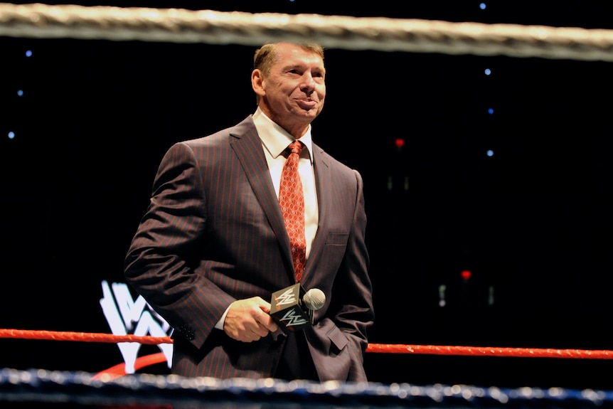 Vince McMahon in a suit and tie holding a microphone inside a wrestling ring with the WWE logo behind him