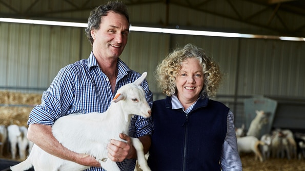 A man and a woman standing in a shed, with the man holding a small white goat.