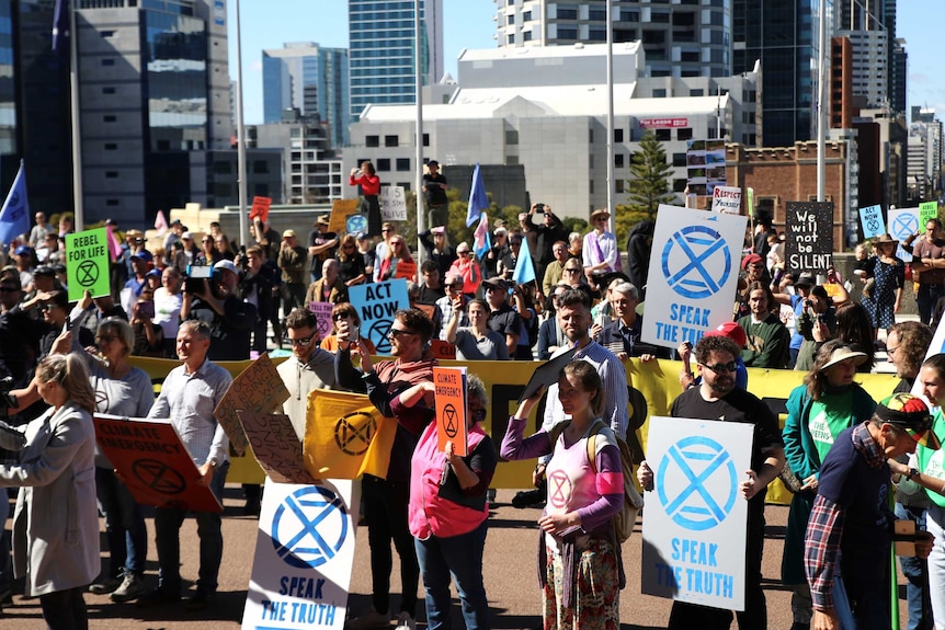 A large group of people, many carrying placards in support of action to tackle climate change, stand with a Perth CBD background