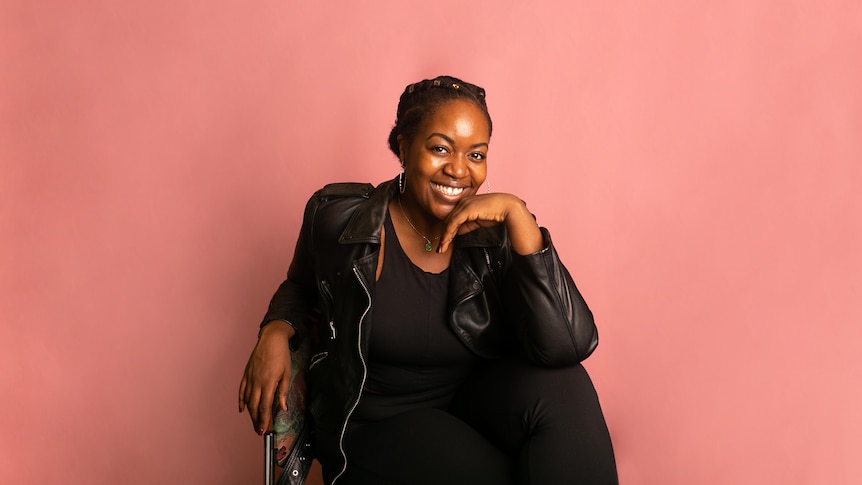 Ruva Ngwenya, a 30-year-old Black woman with braided hair, wears a black leather jacket and hoop earrings before a pink backdrop