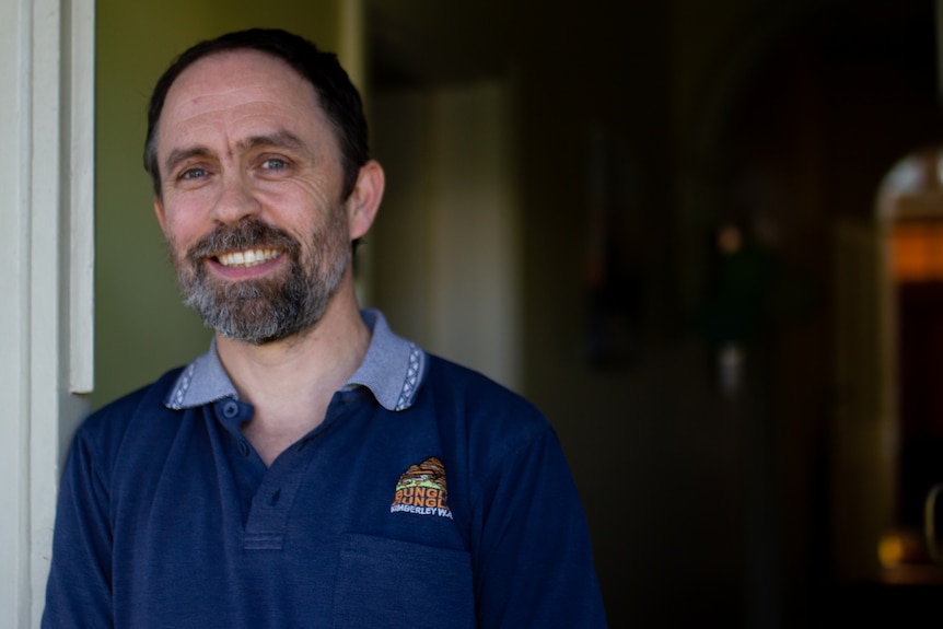 A man with a beard smiles while leaning against his front door frame.