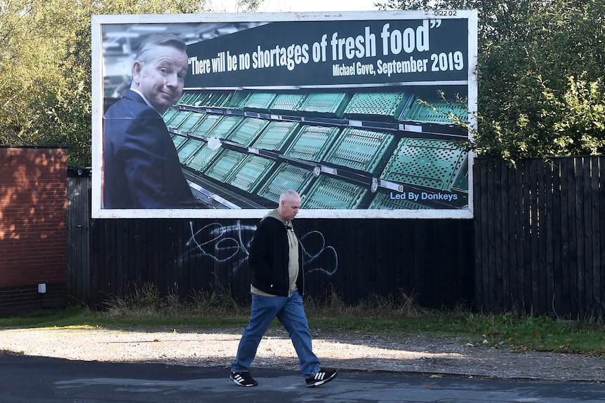 A man walks past a billboard with UK politician Michael Groves, empty shelves, and "there will be no shortages of fresh food".