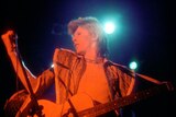 bowie with his head turne,d hand in a fist, performing with his guitar in the ziggy phase