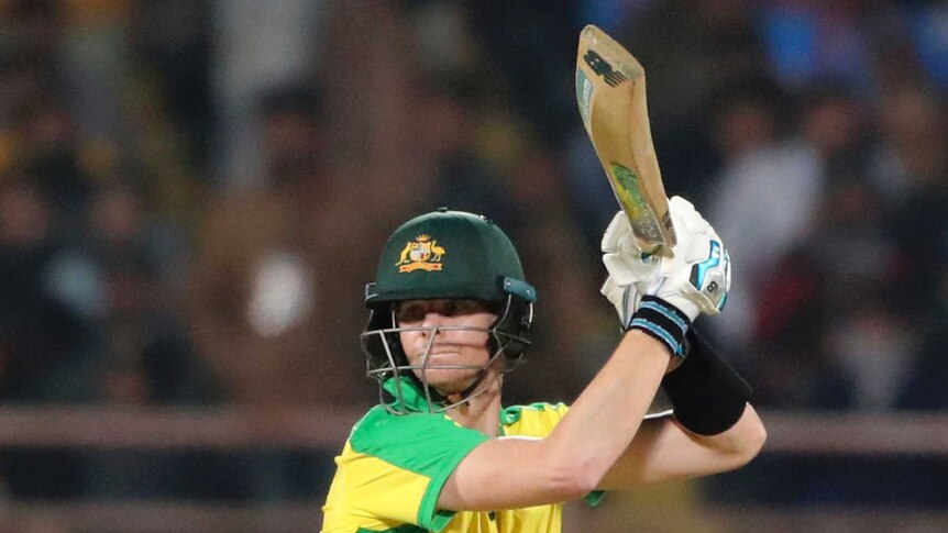 Steve Smith holds his bat above his head and looks behind him wearing yellow cricket kit