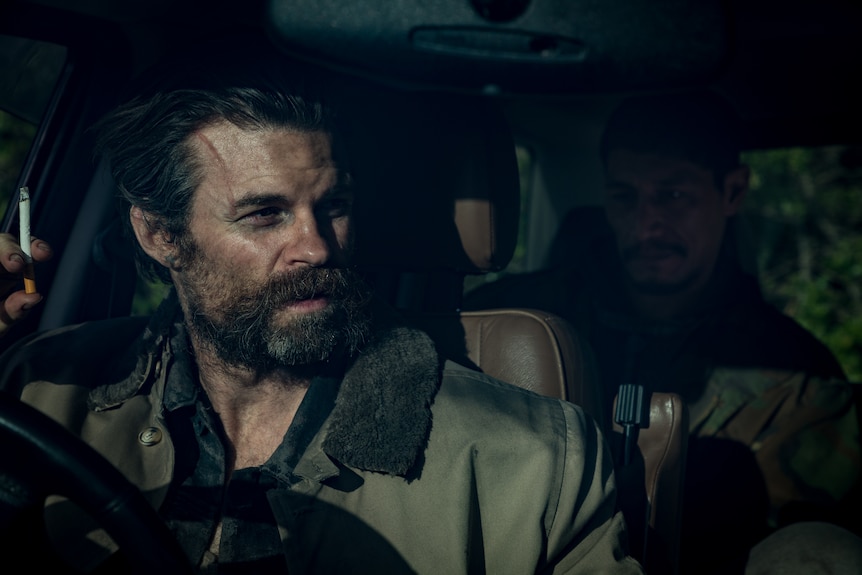 A 40-something man with cropped hair and a messy, thick beard sits in the driver's seat of a darkened car