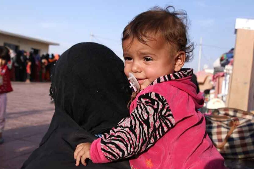 A woman whose face is covered with a black headscarf holds a toddler at a refugee camp