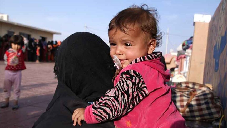 A woman whose face is covered with a black headscarf holds a toddler at a refugee camp