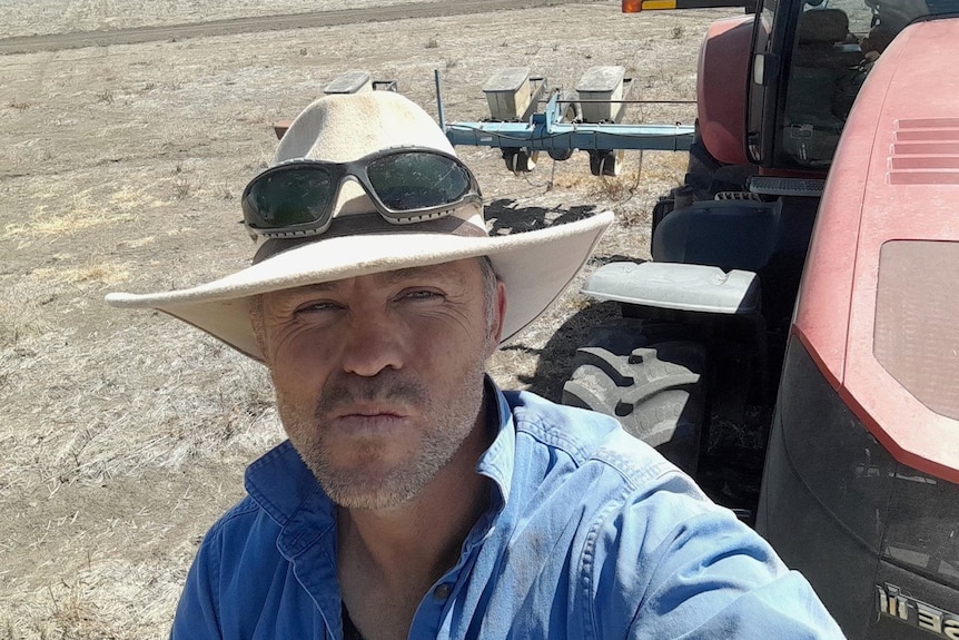 Man in wide brim hat stands in front of a header in a dry paddock