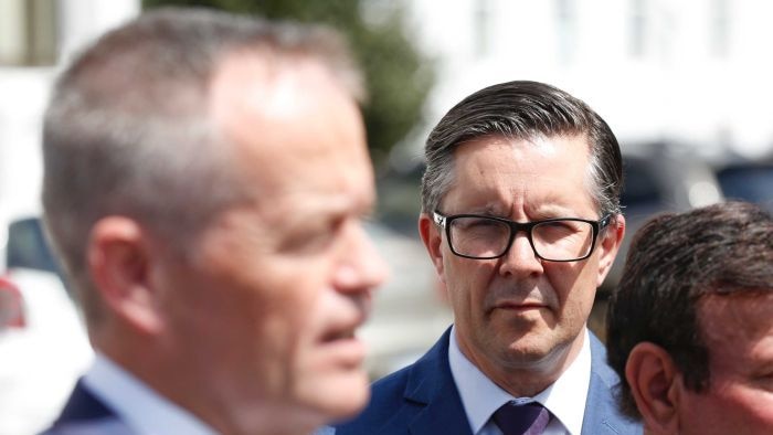 A dark-haired man with glasses looks on at another man who is talking.