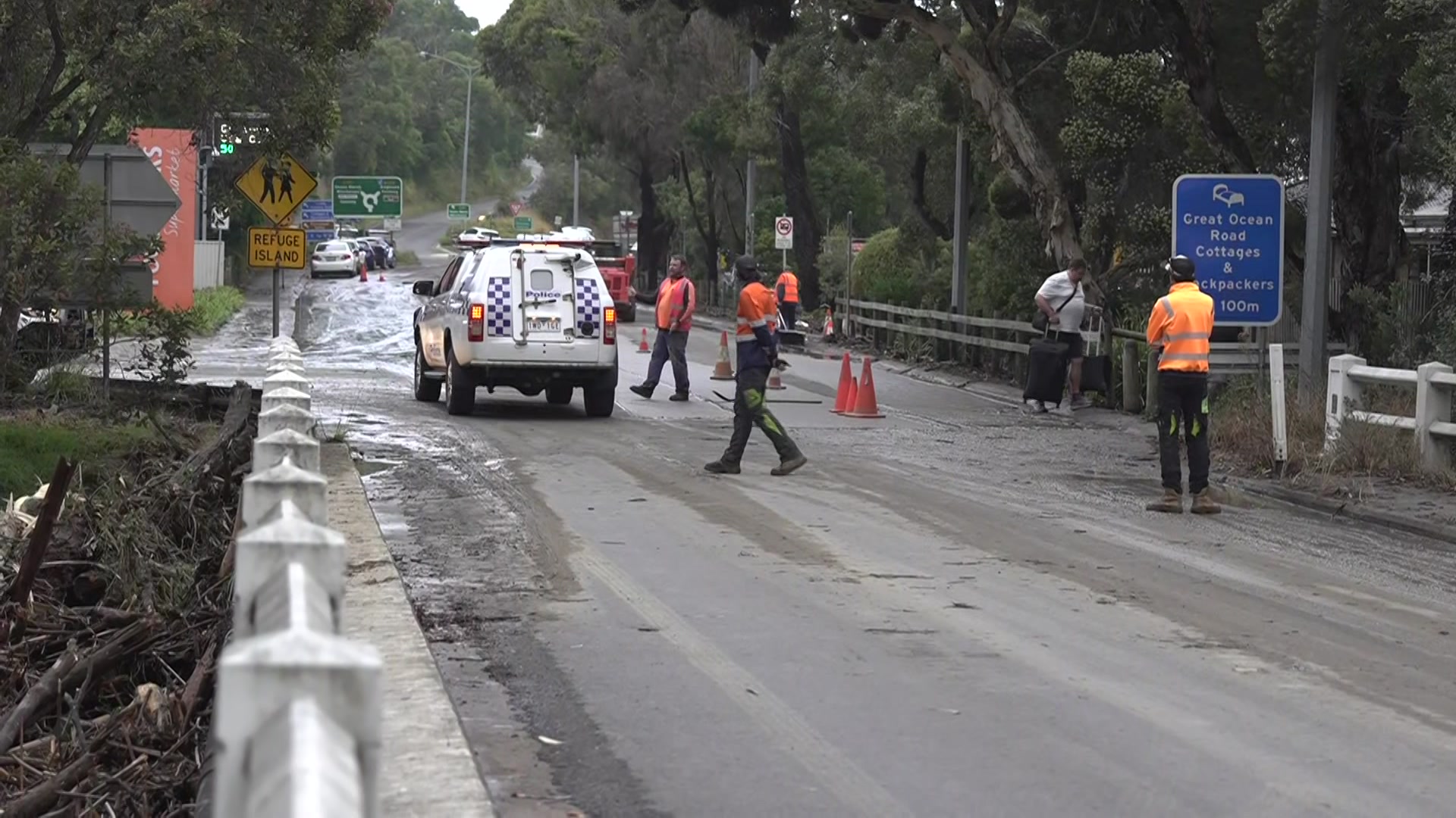 Um carro da polícia e pessoas com camisetas laranja e calças azuis estão em uma estrada coberta de lama.