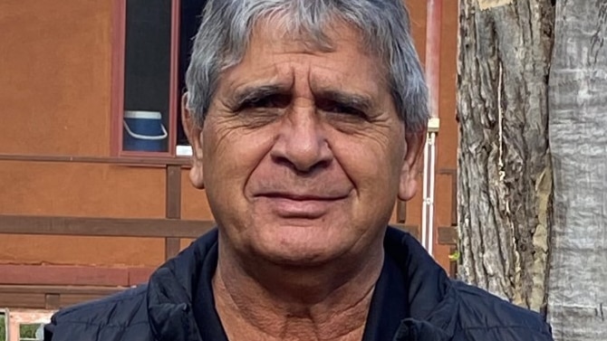 An older Indigenous man with grey hair stands outside in front of a tree.