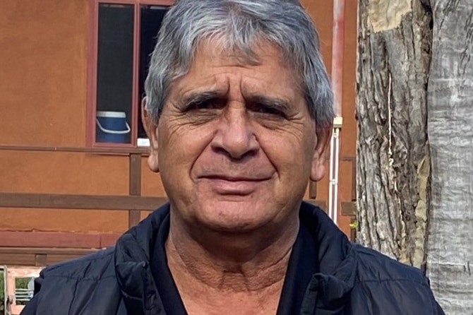 An older Indigenous man with grey hair stands outside in front of a tree.