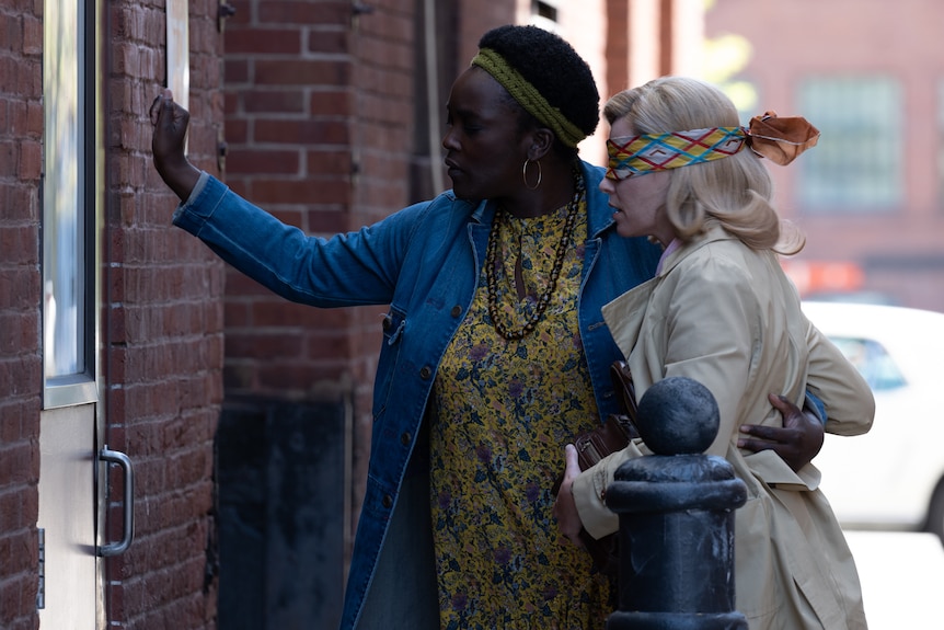 A Black woman knocks at a door. She has her arm around a blindfolded middle-aged woman in a coat