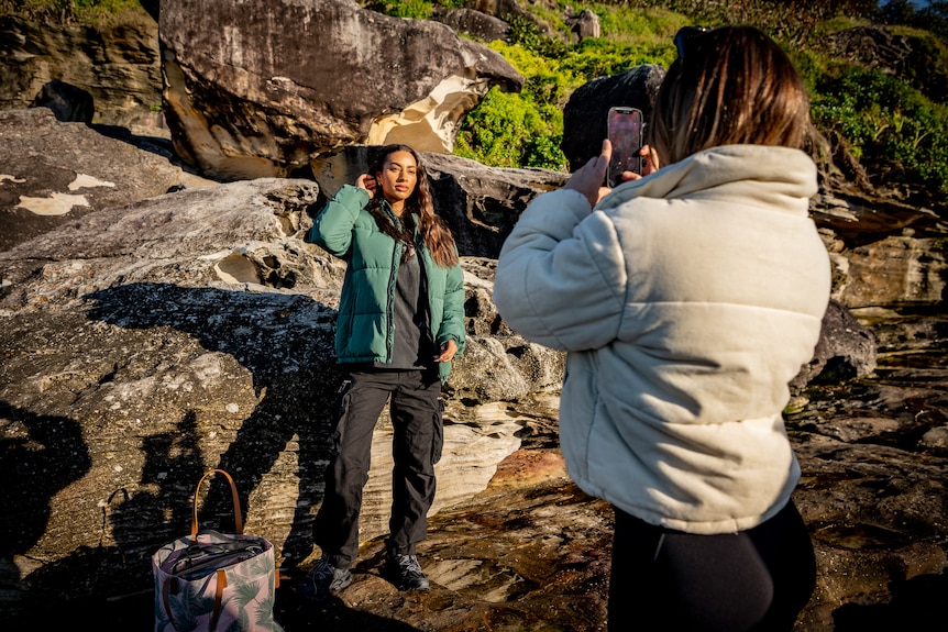 Woman wearing white jacket takes photo of woman in green jacket, with rocks behind her.