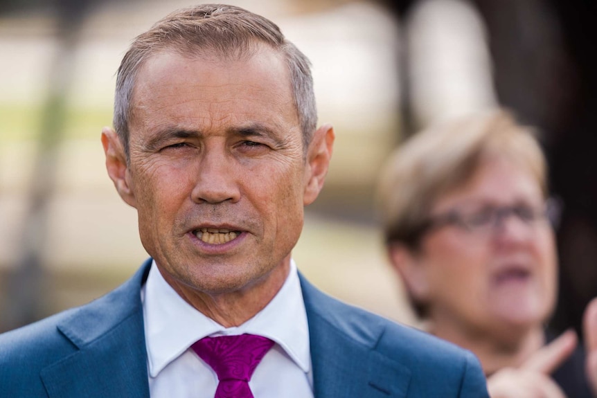 Roger Cook wearing a blue suit and pink tie, with an AUSLAN interpreter in the background.