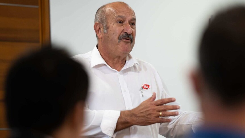 A head and shoulders shot of a man wearing a white business shirt talking in front of a group of people.