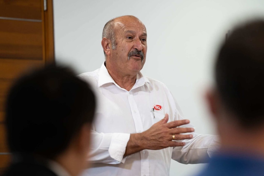 A head and shoulders shot of a man wearing a white business shirt talking in front of a group of people.