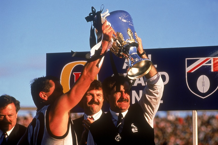 Leigh Matthews and Tony Shaw hold up the 1990 AFL premiership trophy.