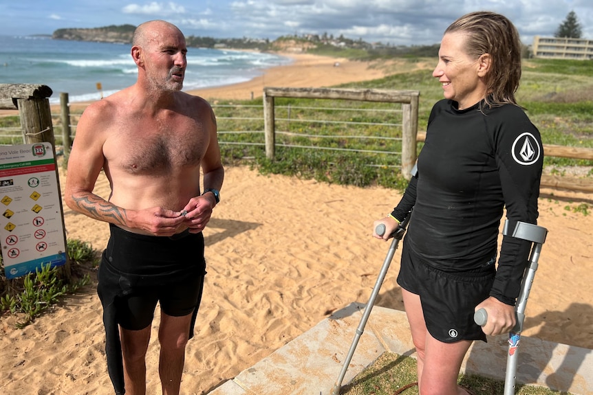Emma Dieters looks at her husband while he speaks. They are standing on the sand at Mona Vale beach, wearing wetsuits.