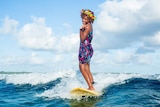 A woman standing up on a surfboard, riding a wave, while wearing flowers in her hair and holding a lollipop.
