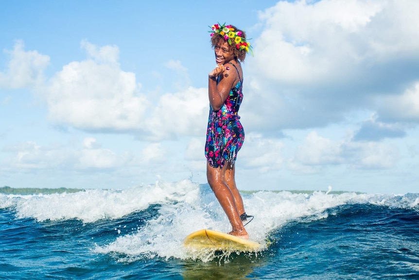 A woman standing up on a surfboard, riding a wave, while wearing flowers in her hair and holding a lollipop.