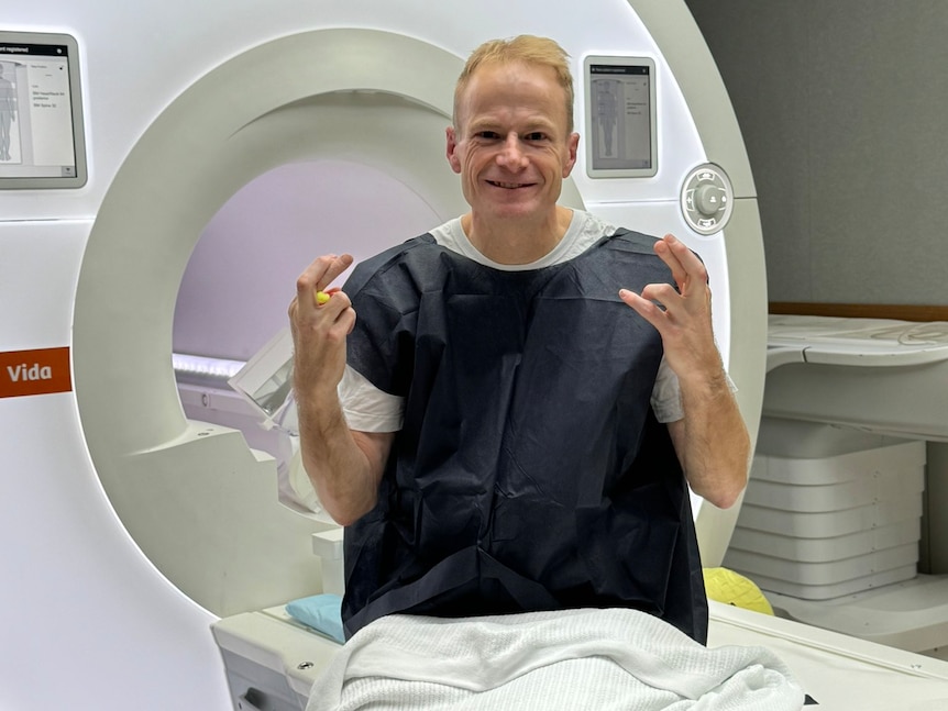 Man in hospital patient gown crosses fingers while sitting next to MRi machine