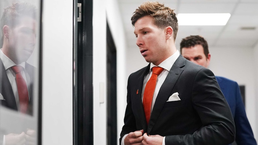 A male AFL player wearing a dark suit, white shirt and orange tie walks through an open door.
