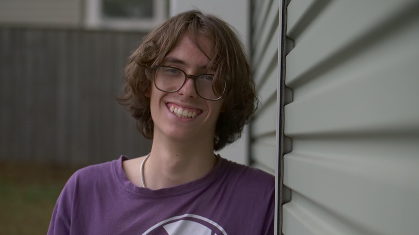 young boy wearing a purple t-shirt and large reading glasses looks into the camera smiling. 