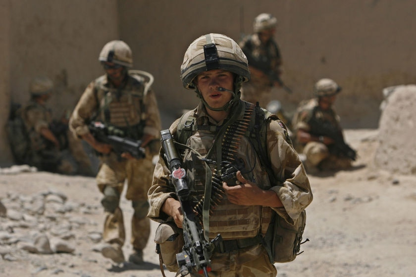 A soldier in battle gear walks across rubble and dust holding a radio and a gun while others sit in shade near a large wall