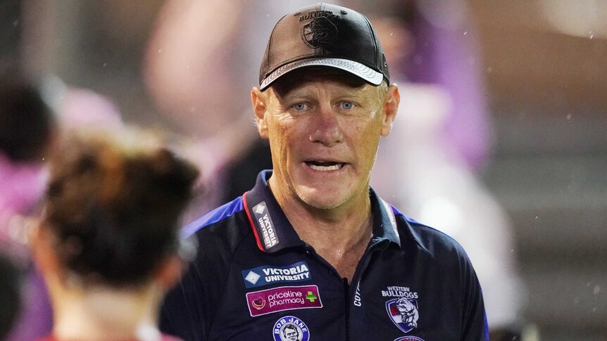 The Western Bulldogs AFLW coach stands talking as he wears a club cap and polo shirt.