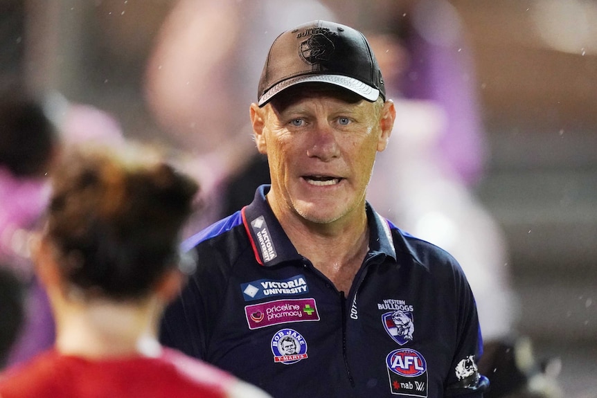 The Western Bulldogs AFLW coach stands talking as he wears a club cap and polo shirt.