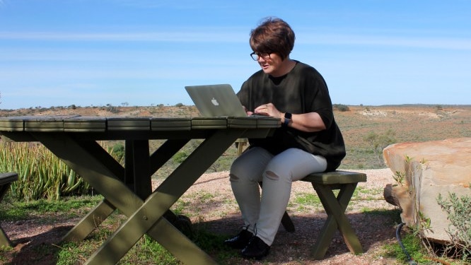 A woman sits at a lap top at a table outside in a bush setting.