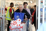 An elderly man in black pushes a shopping trolley through the sliding door of a crowded supermarket.