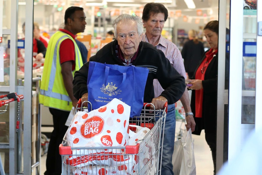 An elderly man in black pushes a shopping trolley through the sliding door of a crowded supermarket.