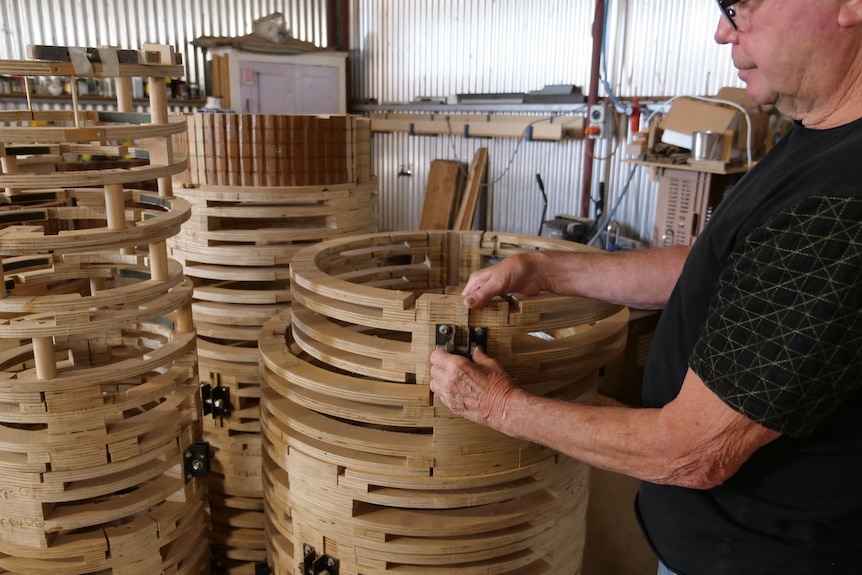 A man wears a dark shirt, standing in a tin shed, he looks at wooden circles piled up